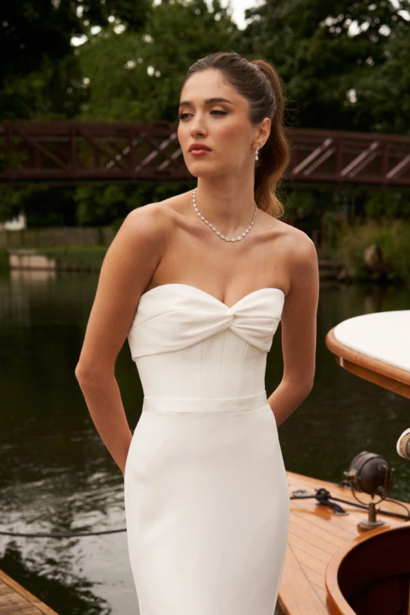 Model wearing a plain fitted wedding dress standing next to a river and boats.